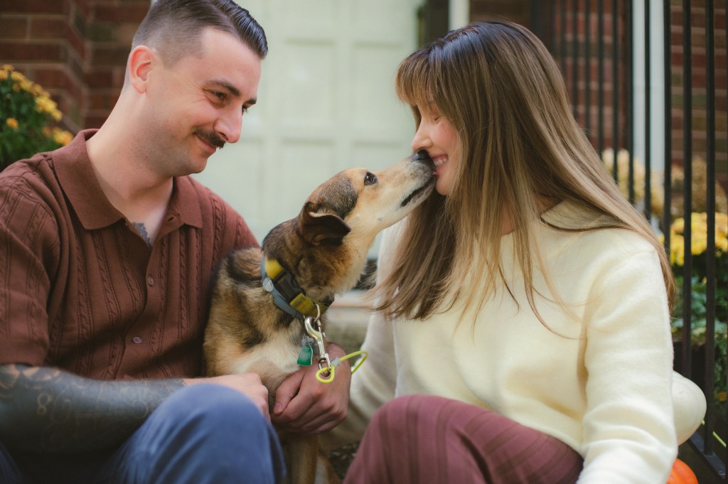 couple looks lovingly at dog while it kisses the woman. 